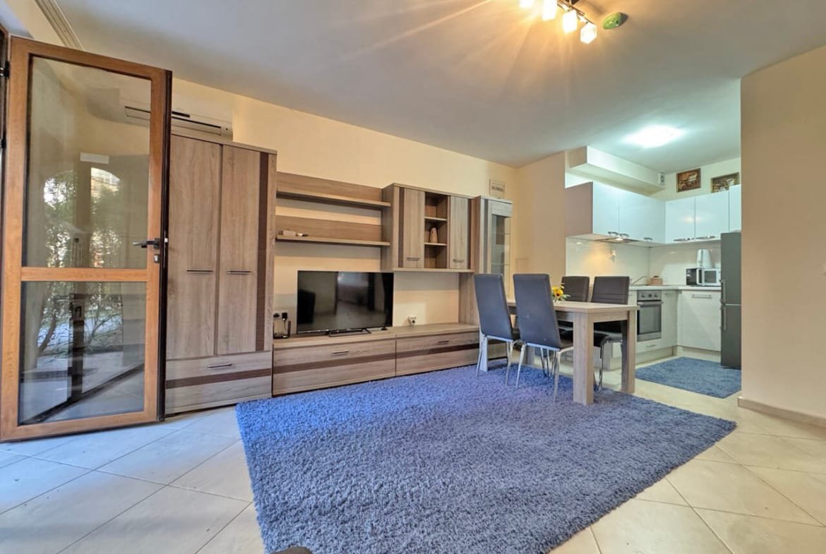 Современная гостиная с телевизором, столом на шесть человек и кухонной зоной, оформленная в светлых тонах.