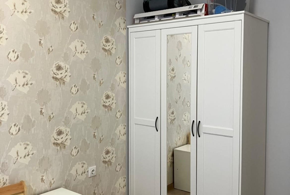 Современная спальня с белым шкафом, прикроватной тумбочкой и цветочными обоями, подходящая для покупки недвижимости в Болгарии.