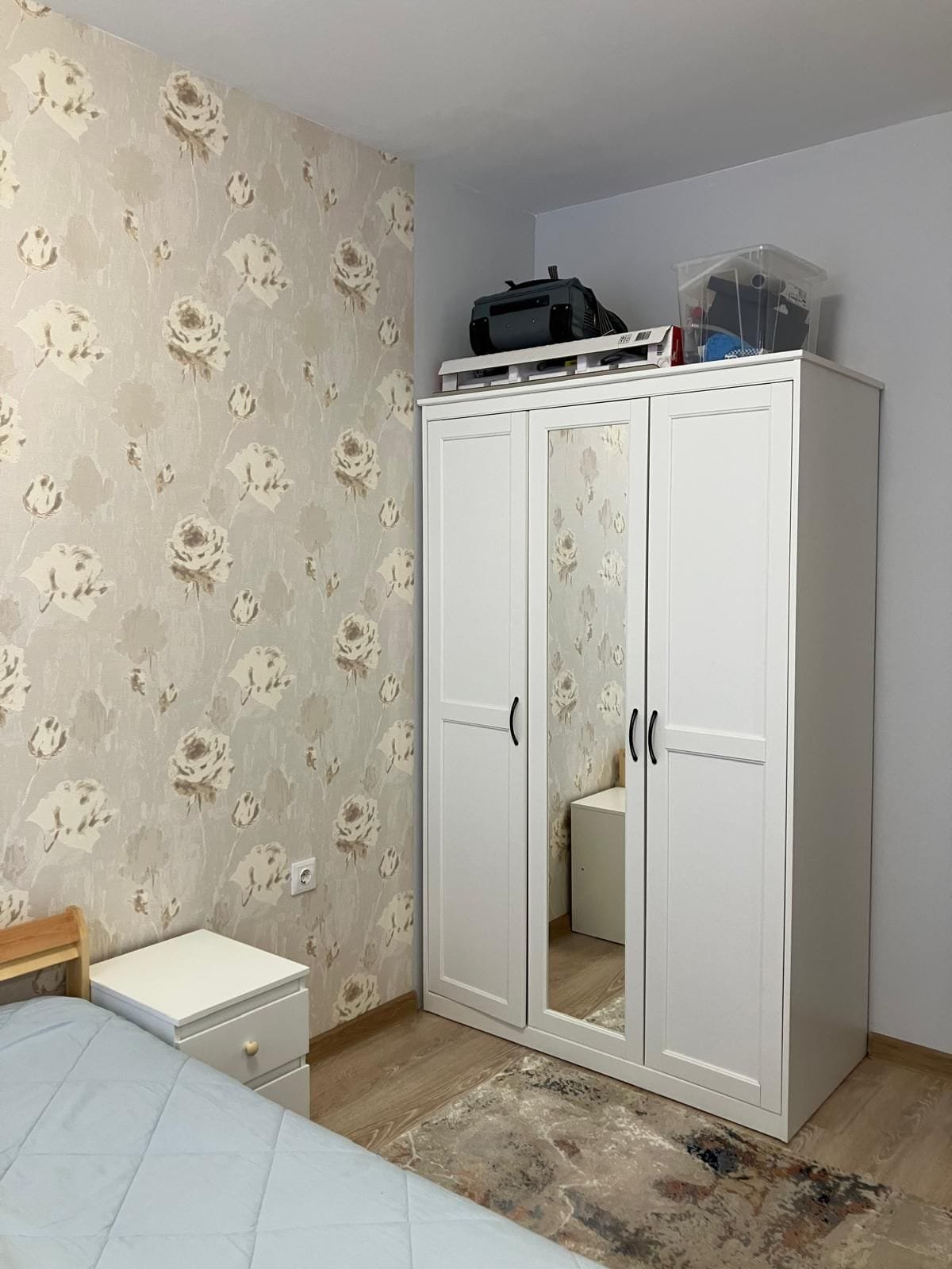 Современная спальня с белым шкафом, прикроватной тумбочкой и цветочными обоями, подходящая для покупки недвижимости в Болгарии.