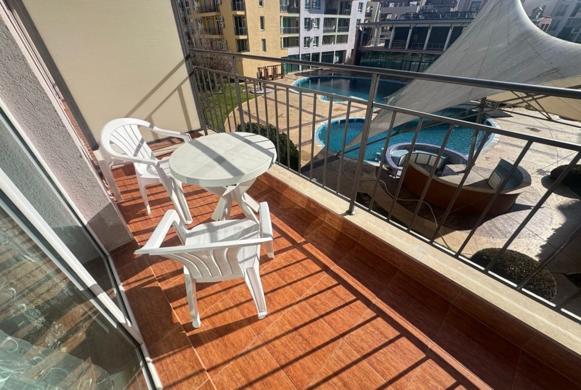 Балкон с двумя белыми креслами и столом, рядом с солнечным бассейном и современными зданиями, подходит для отдыха и покупки недвижимости в Болгарии.