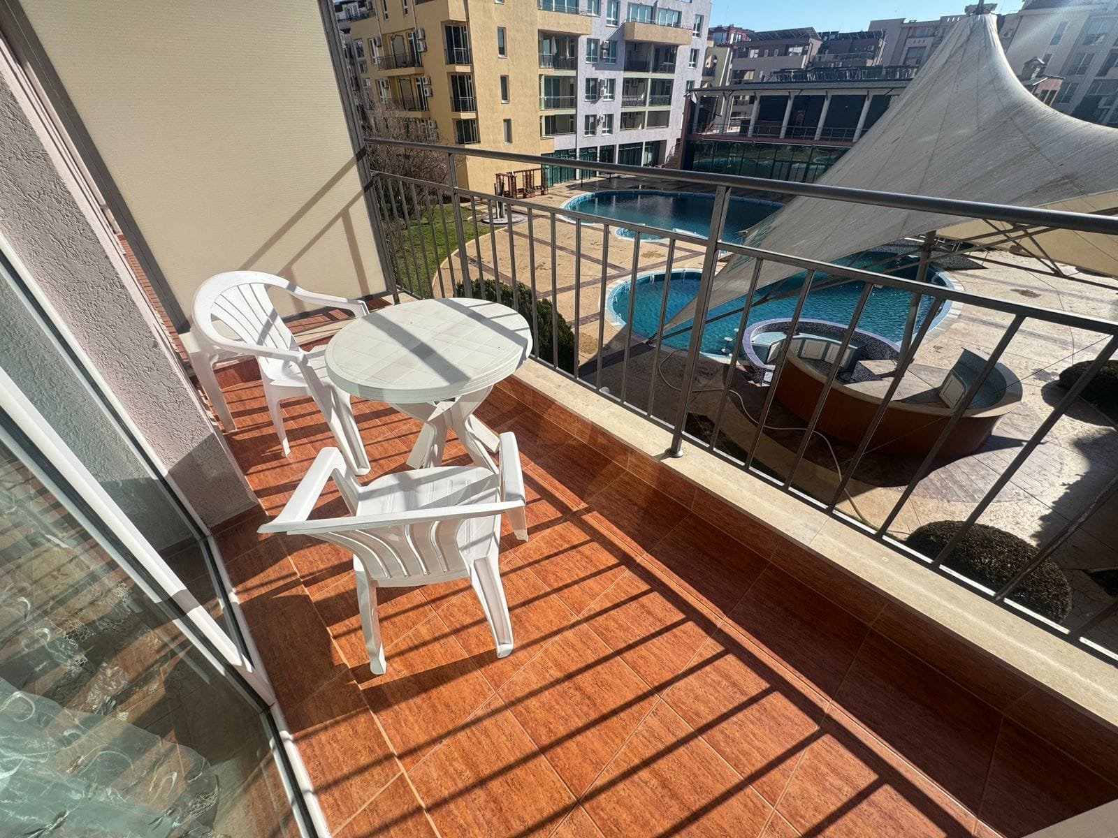 Балкон с двумя белыми креслами и столом, рядом с солнечным бассейном и современными зданиями, подходит для отдыха и покупки недвижимости в Болгарии.