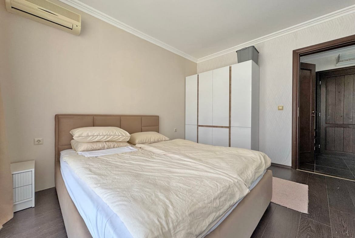 Современная спальня с двуспальной кроватью, светлыми стенами и белым шкафом, пол из темного дерева.