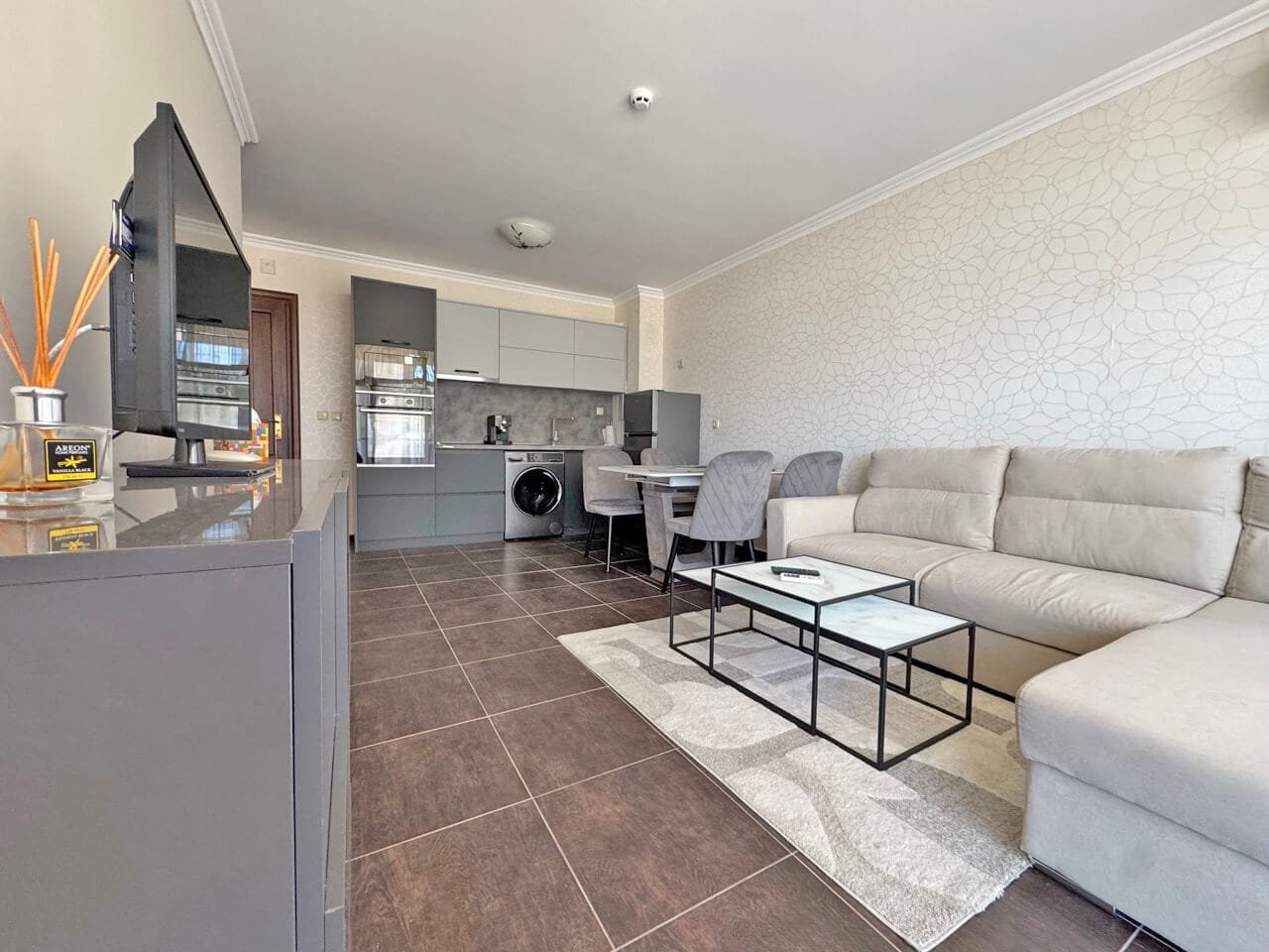 Современный интерьер квартиры: уютная гостиная с кухней Современный интерьер квартиры с серым диваном, стеклянным столом и кухонной зоной, оформленной в светлых тонах.