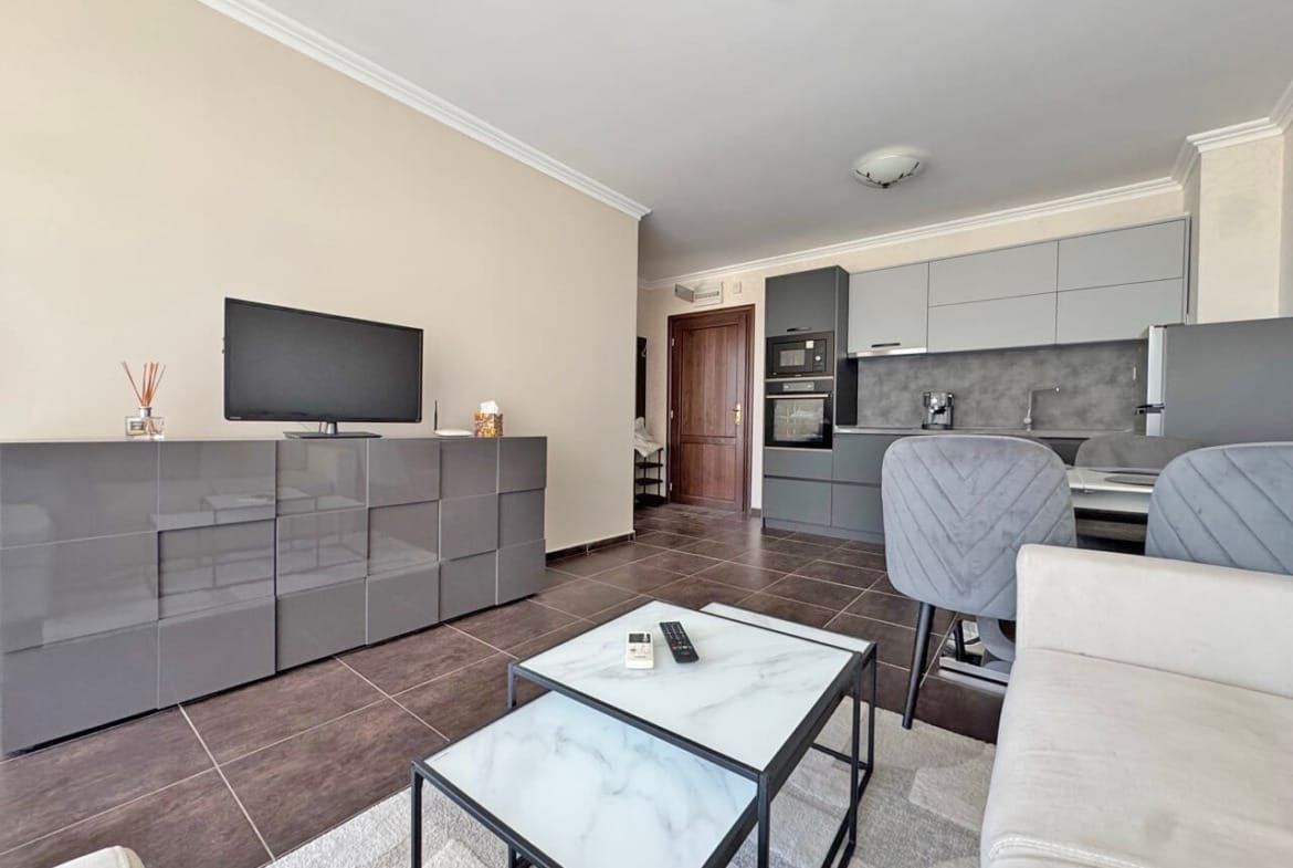 Современный интерьер квартиры с серой мебелью, телевизором, кухней и светлым диваном в зонированом пространстве.