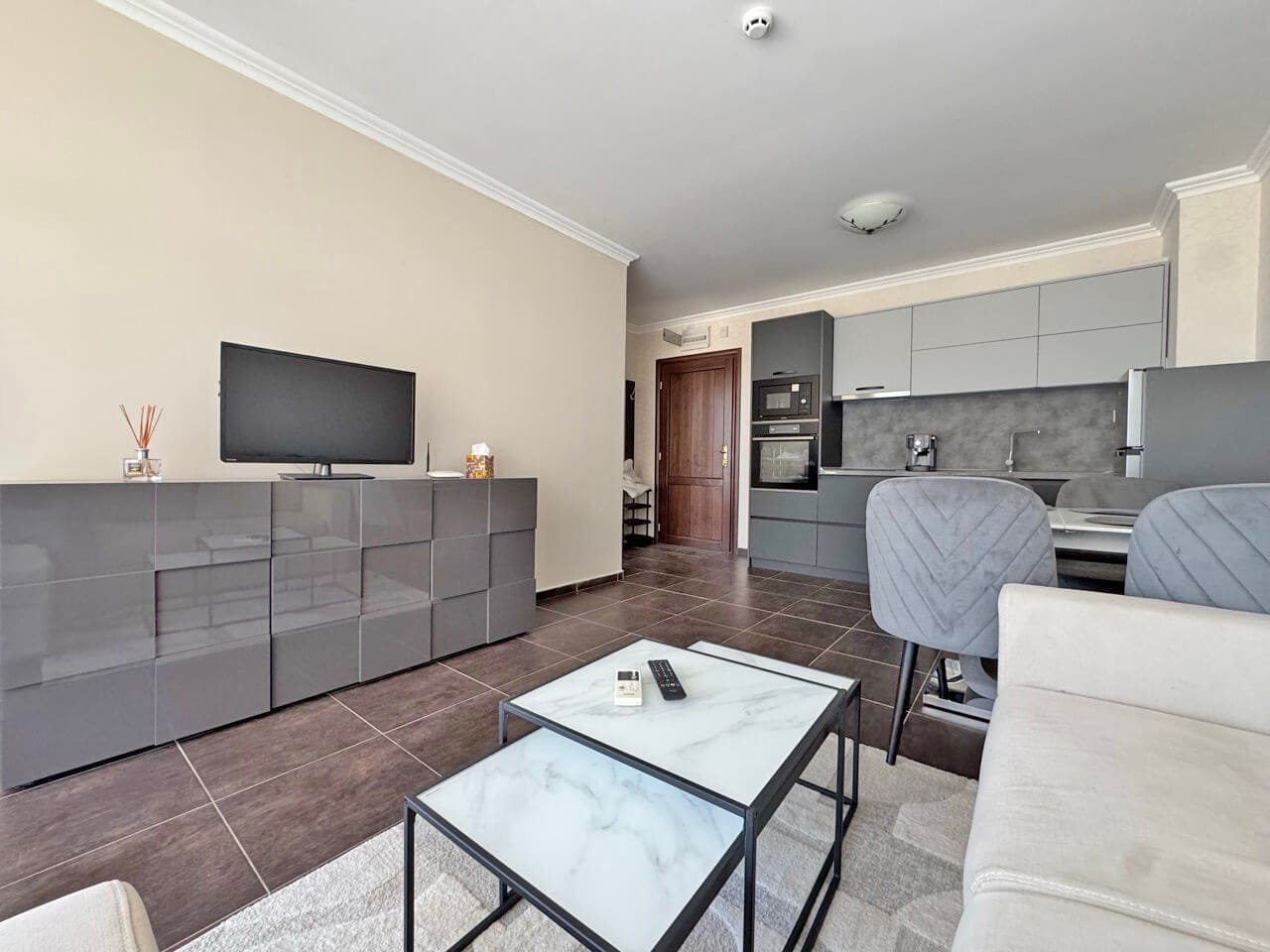 Современный интерьер квартиры: стильная гостиная и кухня Современный интерьер квартиры с серой мебелью, телевизором, кухней и светлым диваном в зонированом пространстве.