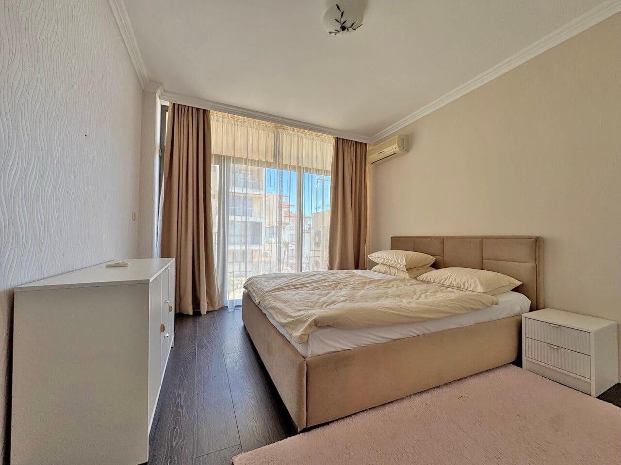 Современная спальня с большим окном и уютной атмосферой Светлая спальня с кроватью, белыми подушками и нежными шторами, окна выходят на улицу, уютный интерьер.