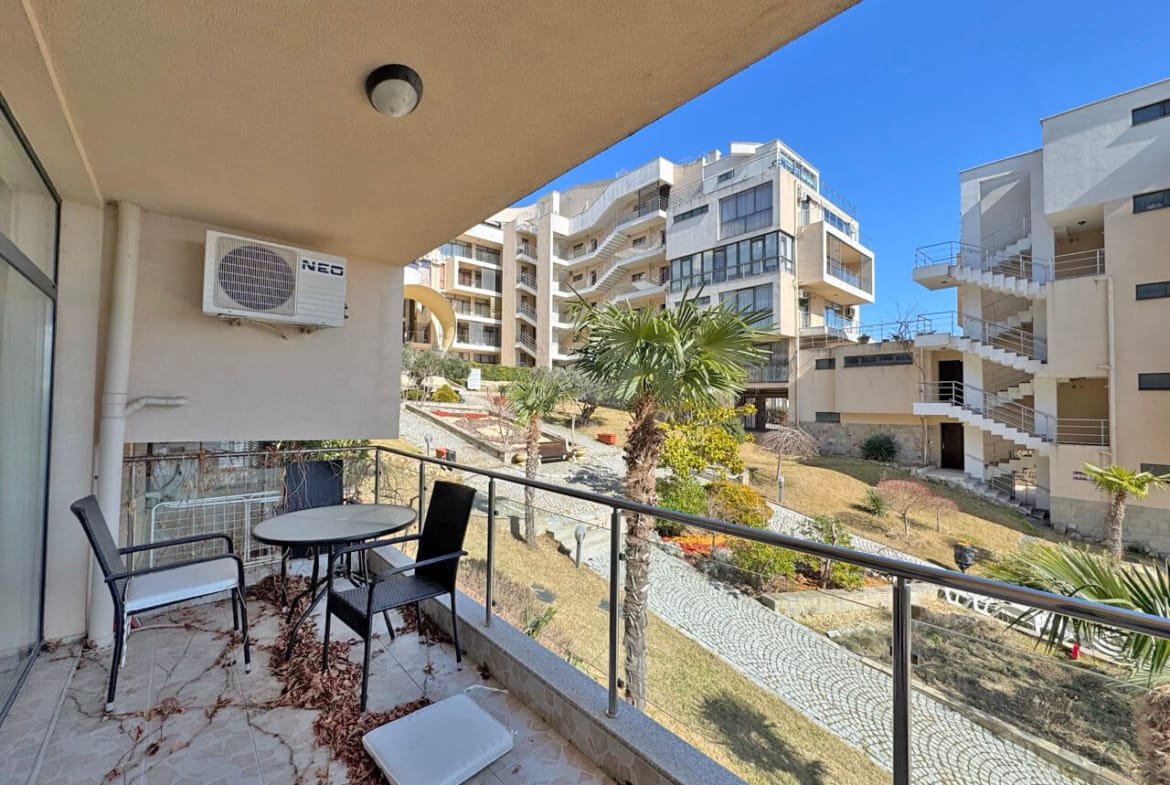 Б Balcony с двумя стульями и столом, вид на зелёный двор и современные здания под чистым небом.