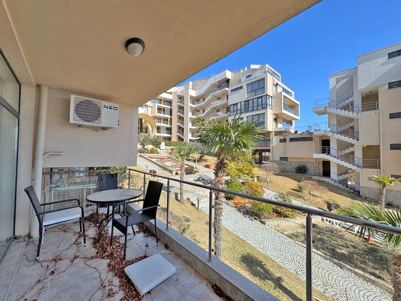 Современный балкон с видом на уютный жилой комплекс Б Balcony с двумя стульями и столом, вид на зелёный двор и современные здания под чистым небом.