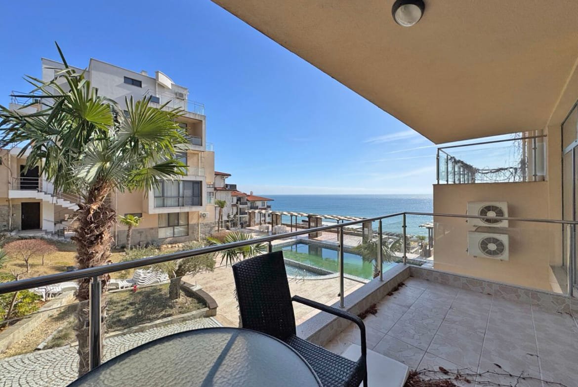 Вид из балкона на море и жилые комплексы, с пальмами и бассейном на переднем плане. Яркое солнечное небо.