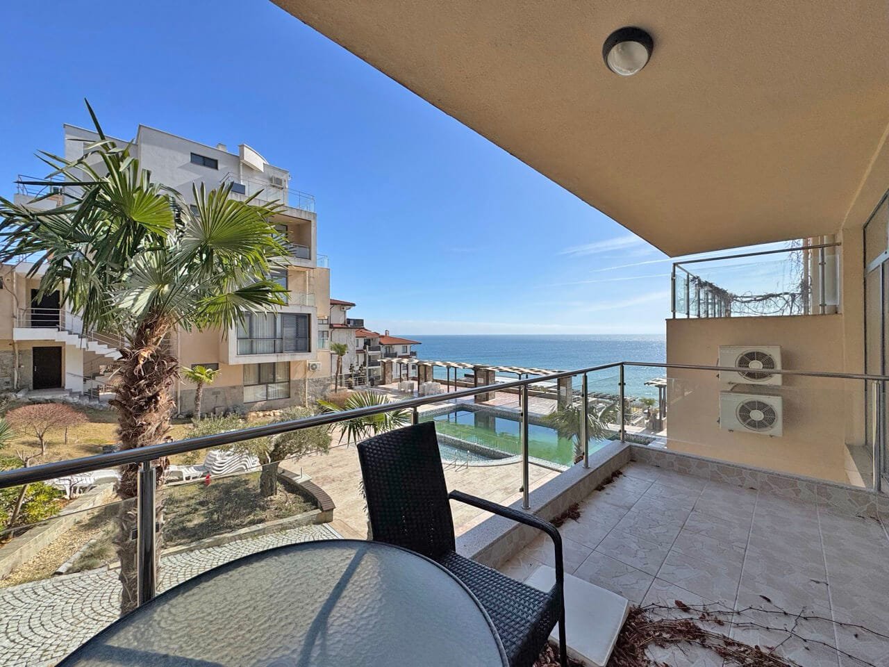 Светлая терраса с видом на море и жилые комплексы у пляжа Вид из балкона на море и жилые комплексы, с пальмами и бассейном на переднем плане. Яркое солнечное небо.