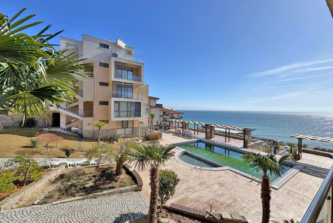 Современный жилой комплекс у моря с бассейном, пальмами и каменной дорожкой, на фоне синего неба и спокойного океана.