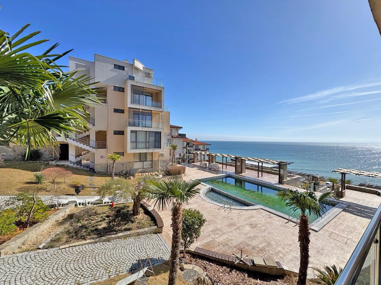 Курортный отель с бассейном и видом на море. Современный жилой комплекс у моря с бассейном, пальмами и каменной дорожкой, на фоне синего неба и спокойного океана.