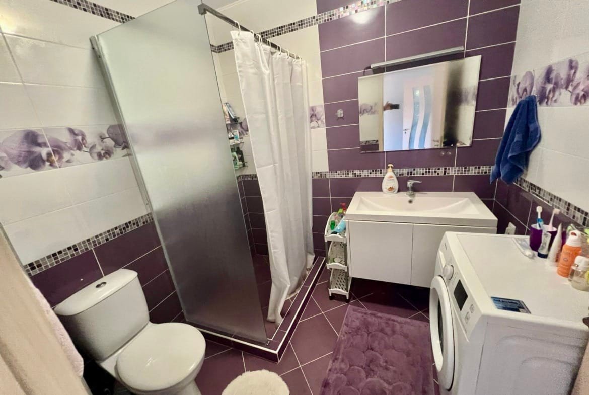 Современная ванная комната с душевой кабиной, умывальником и стиральной машиной, оформленная в фиолетовых и белых тонах.