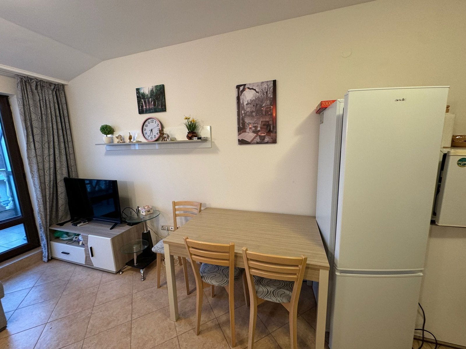 Уютная кухня-студия с телевизором и обеденным столом Современный интерьер квартиры с кухонным столом, холодильником и телевизором, подходит для жизни и аренды в Болгарии.