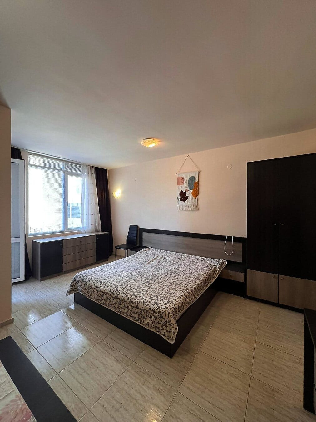 Современная спальня с двуспальной кроватью, комодом и шкафом, окна с видом на улицу, светлые тона интерьера.