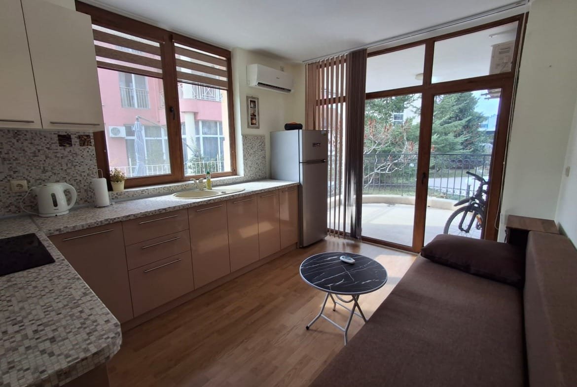 Современная кухня с открытой планировкой, холодильником, обеденным столом и диваном, окнами с видом на балкон и зелень.