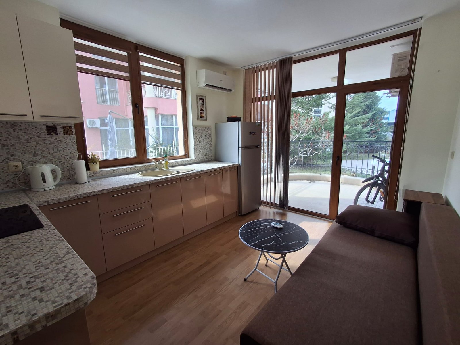 Современная кухня с открытой планировкой, холодильником, обеденным столом и диваном, окнами с видом на балкон и зелень.