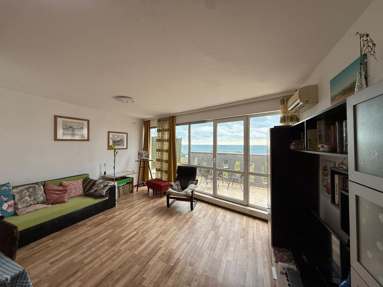 Современная гостиная с зелёным диваном и большим окном, предлагающим вид на море, подходит для жизни или аренды в Болгарии.