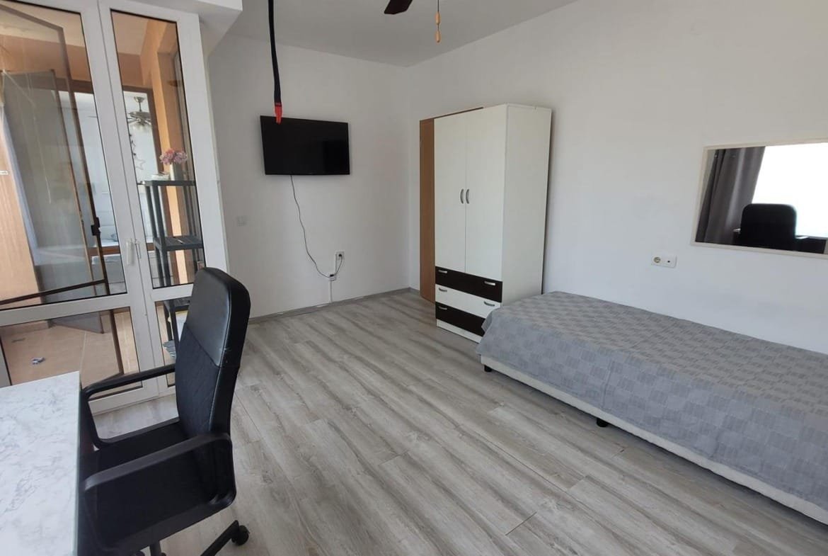 Светлая комната с кроватью, столом и телевизором, окна выходят на балкон, подходящая для покупки недвижимости в Болгарии.