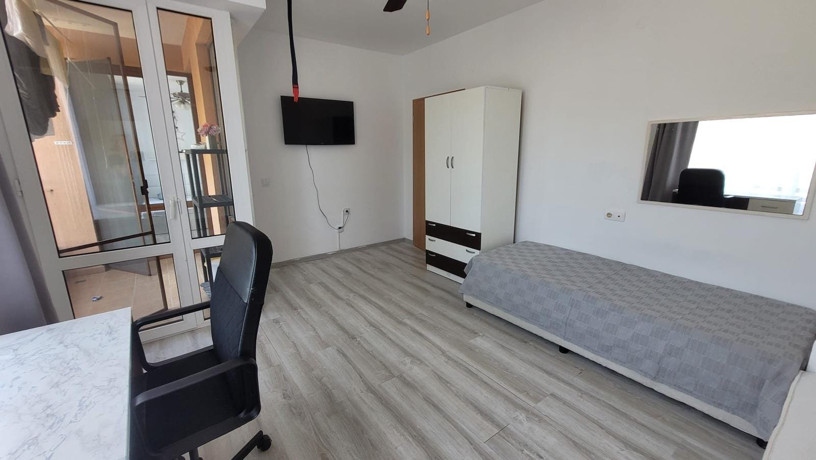 Светлая комната с кроватью, столом и телевизором, окна выходят на балкон, подходящая для покупки недвижимости в Болгарии.