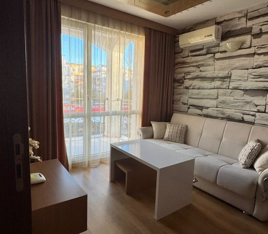 Современная гостиная с мягким диваном, столиком и большими окнами, оформленная в светлых тонах для покупки недвижимости в Болгарии.