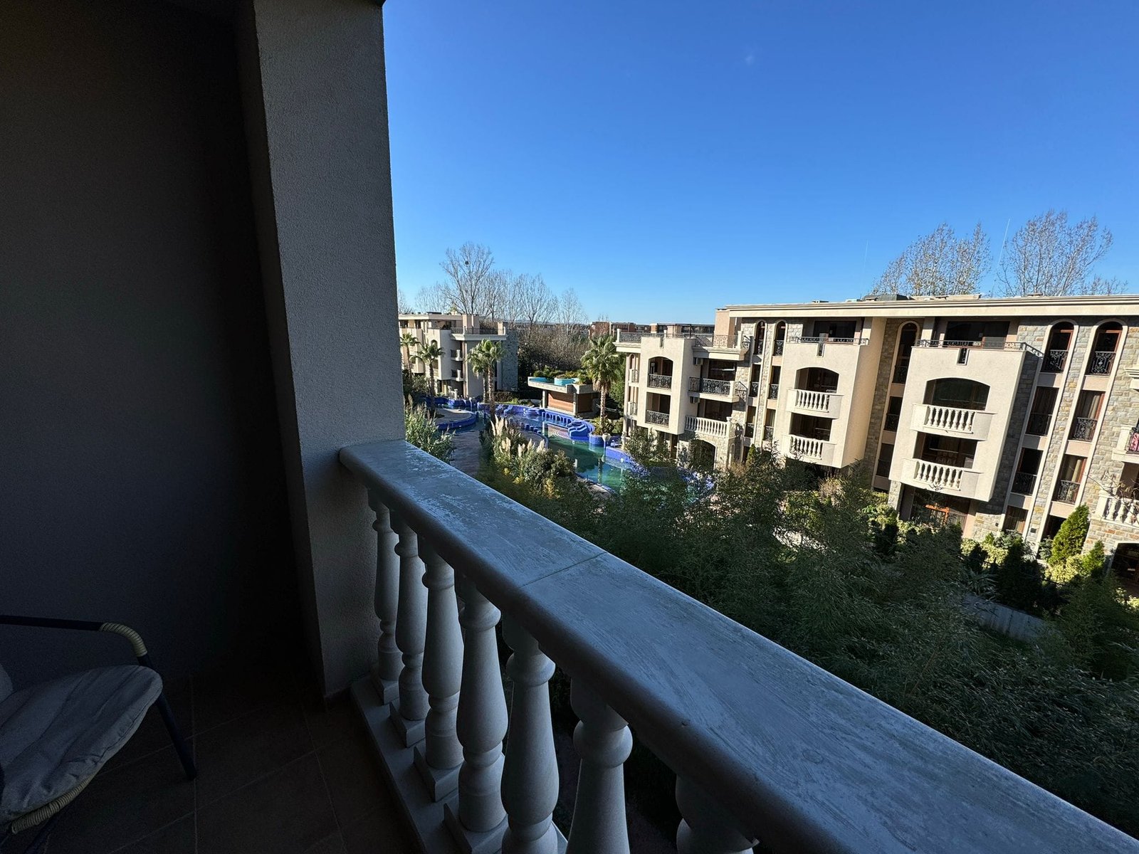 Вид с балкона на жилой комплекс и бассейн под ясным небом Балкон с видом на жилой комплекс в Болгарии, бассейн и зеленые зоны, солнечное небо и уютная атмосфера.