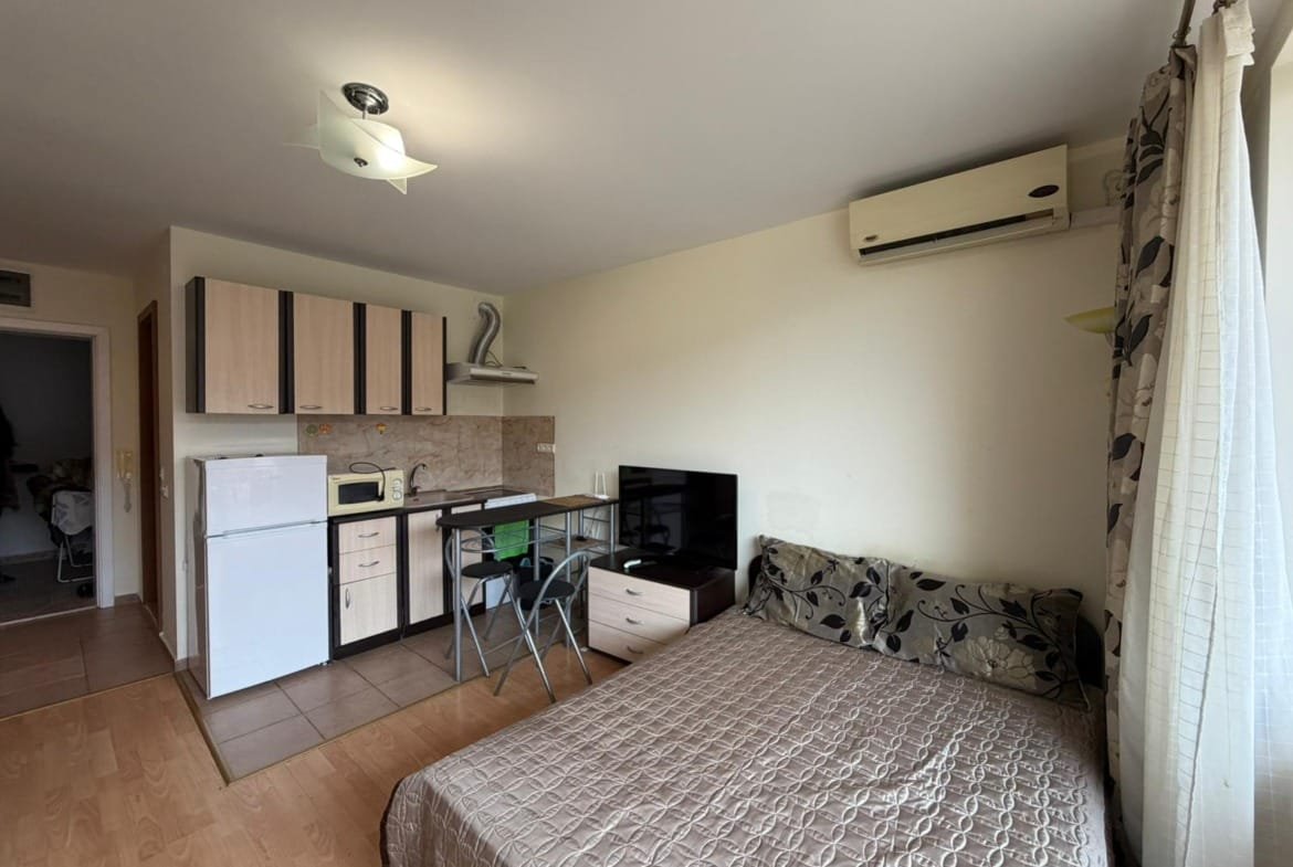 Современная студия с кухонным уголком, обеденным столом, телевизором и диваном, идеальна для покупки недвижимости в Болгарии.