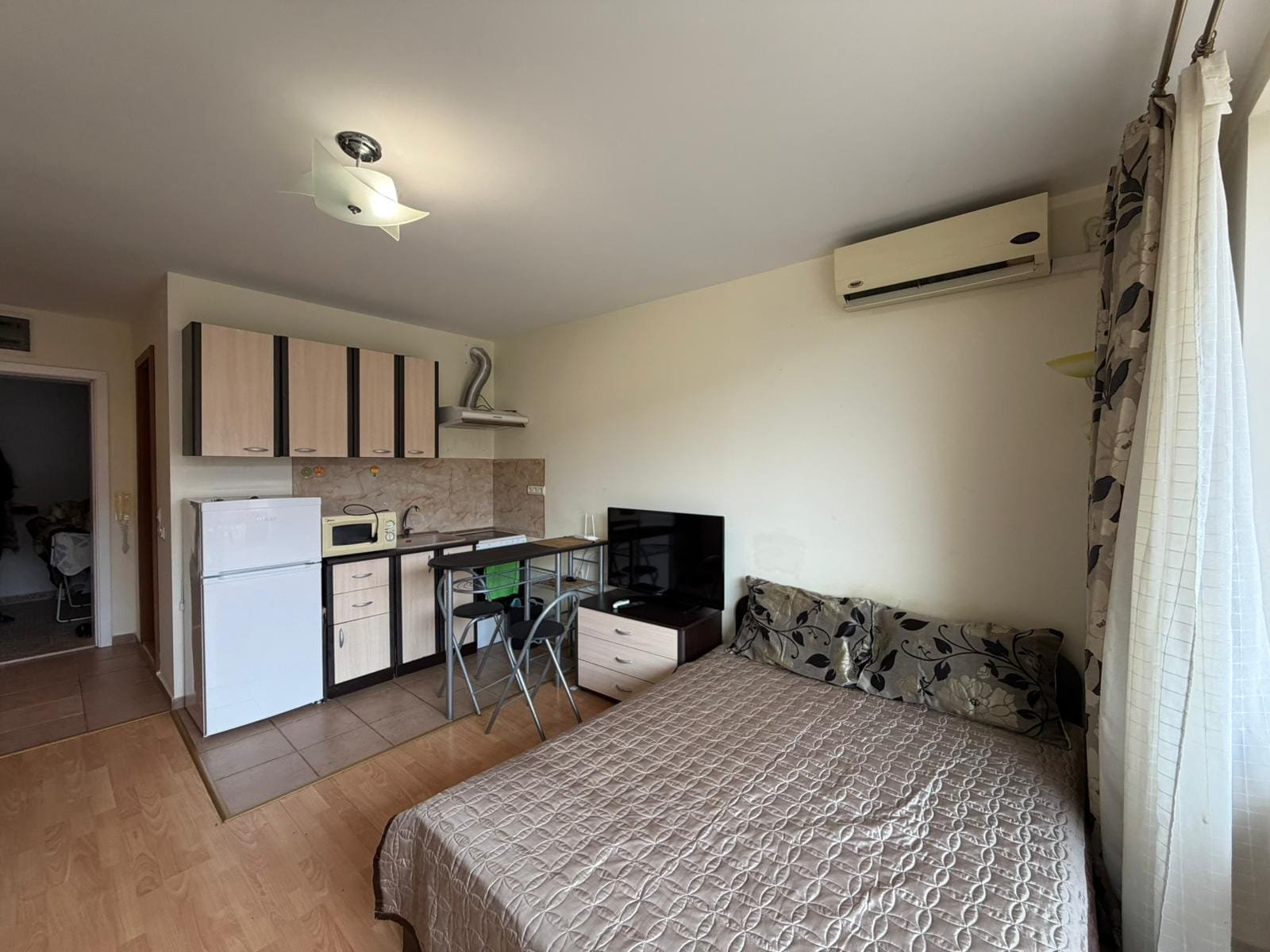 Современная студия с кухней и спальней в светлых тонах Современная студия с кухонным уголком, обеденным столом, телевизором и диваном, идеальна для покупки недвижимости в Болгарии.