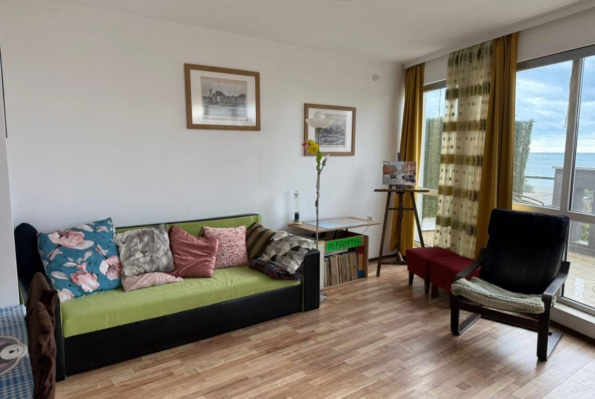 Светлая комната с диваном, столом и стулом, окна с видом на море, уютная атмосфера для отдыха на побережье Болгарии.