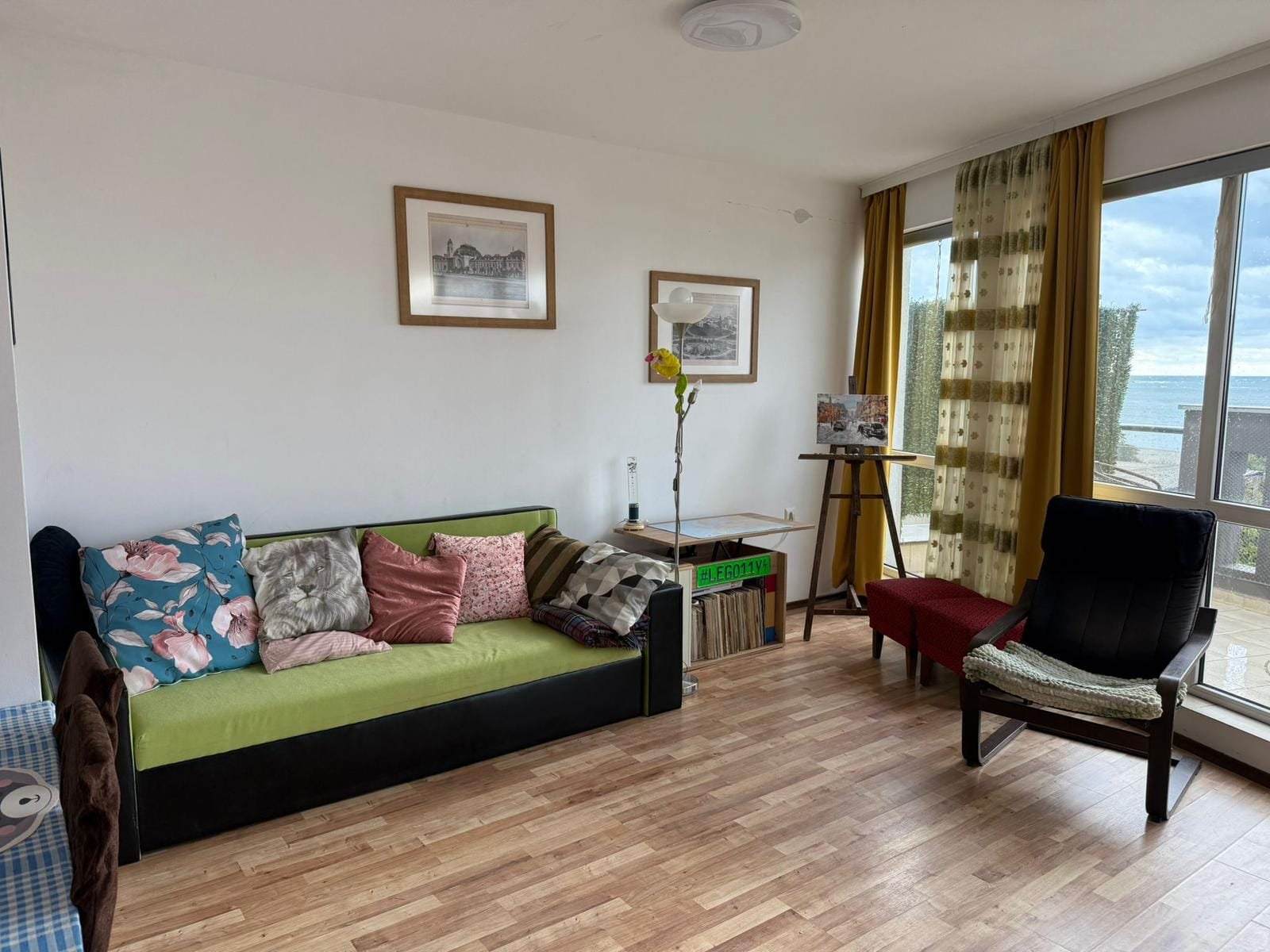 Светлая комната с диваном, столом и стулом, окна с видом на море, уютная атмосфера для отдыха на побережье Болгарии.