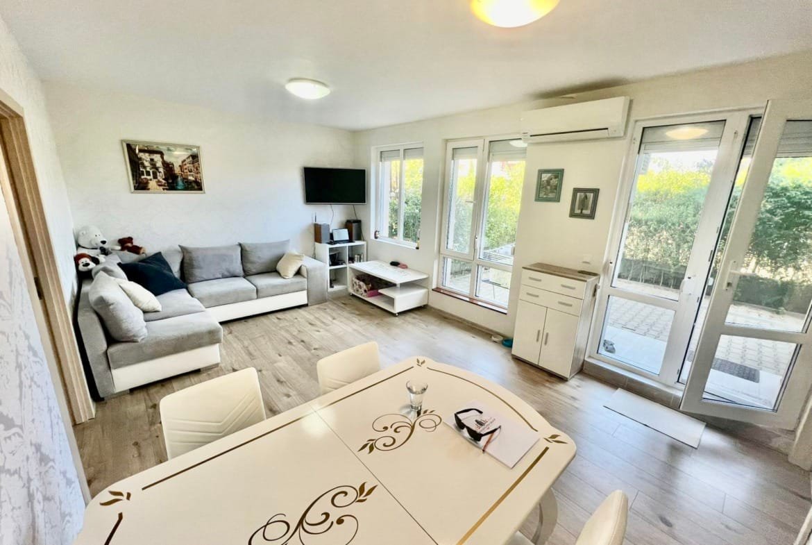 Современная квартира с уютной гостиной, обеденным столом и большими окнами, выходящими на зелёный двор в Болгарии.