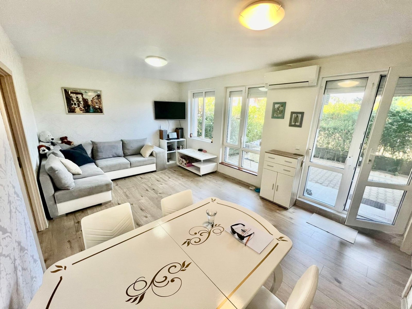 Современная квартира с уютной гостиной, обеденным столом и большими окнами, выходящими на зелёный двор в Болгарии.