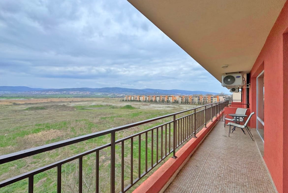 Балкон с креслом, вид на пустырь и здания на горизонте под облачным небом.