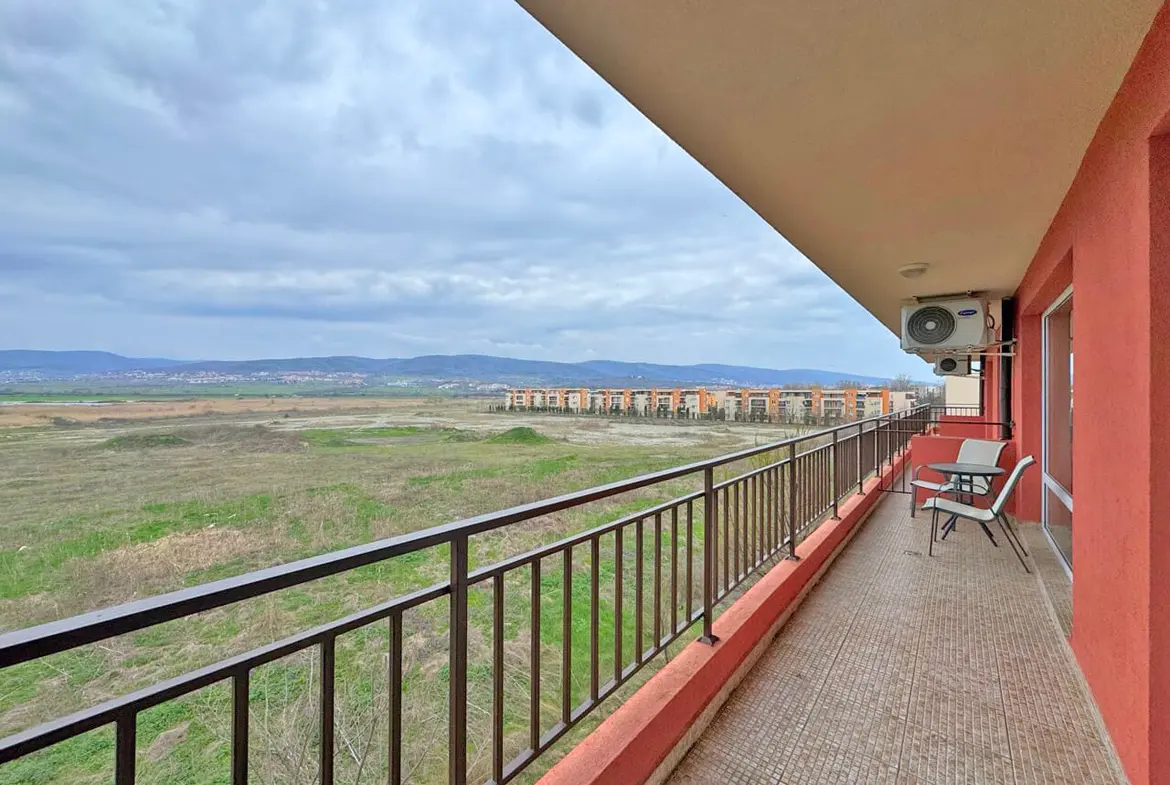 Балкон с креслом, вид на пустырь и здания на горизонте под облачным небом.