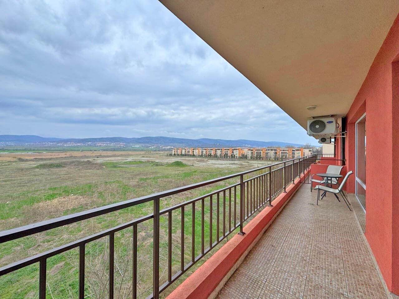 Балкон с креслом, вид на пустырь и здания на горизонте под облачным небом.