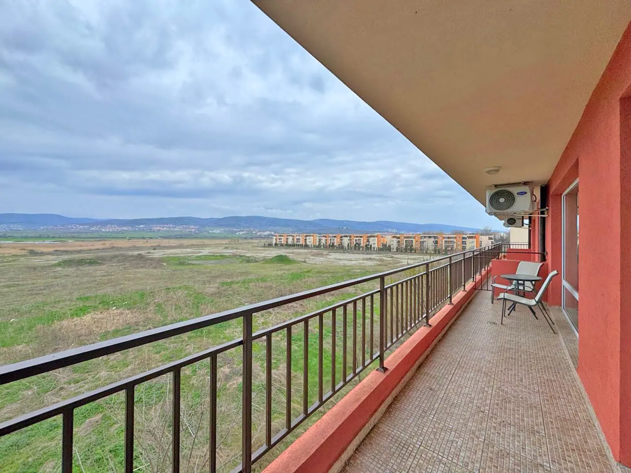 Балкон с креслом, вид на пустырь и здания на горизонте под облачным небом.