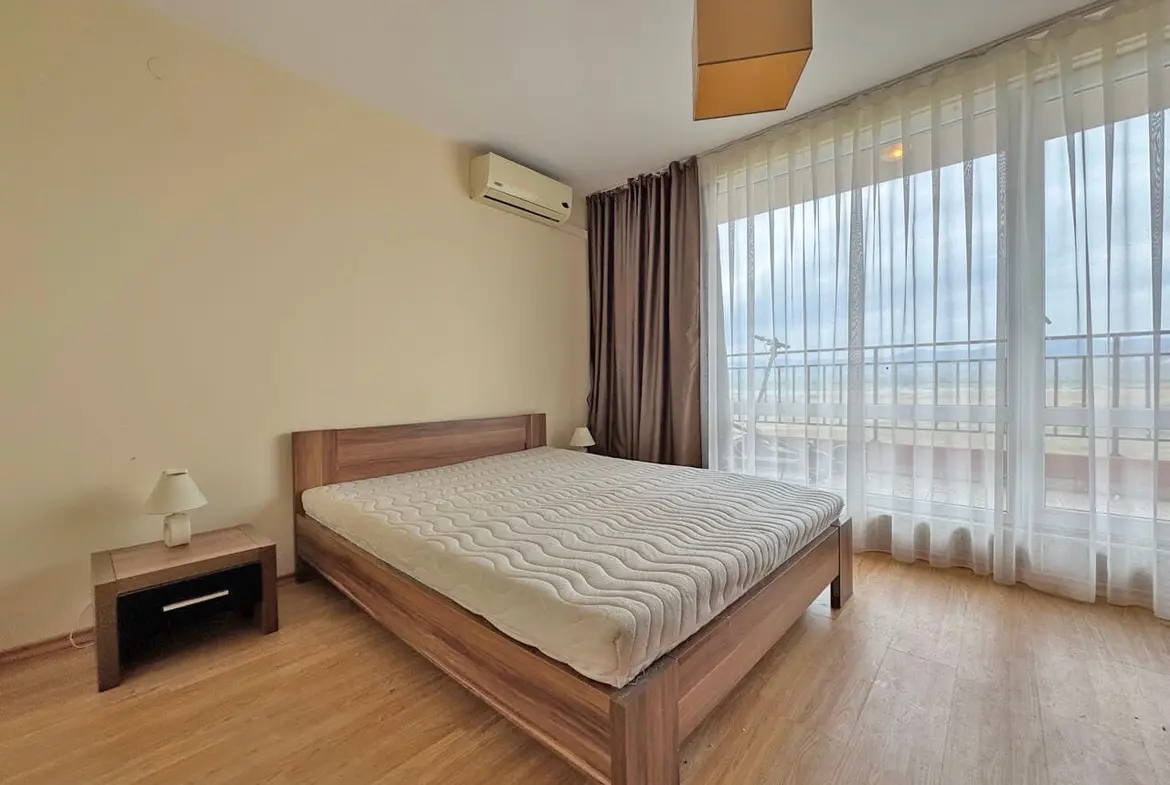 Современная светлая спальня с деревянной кроватью, прикроватной тумбочкой и большими окнами, выходящими на балкон.