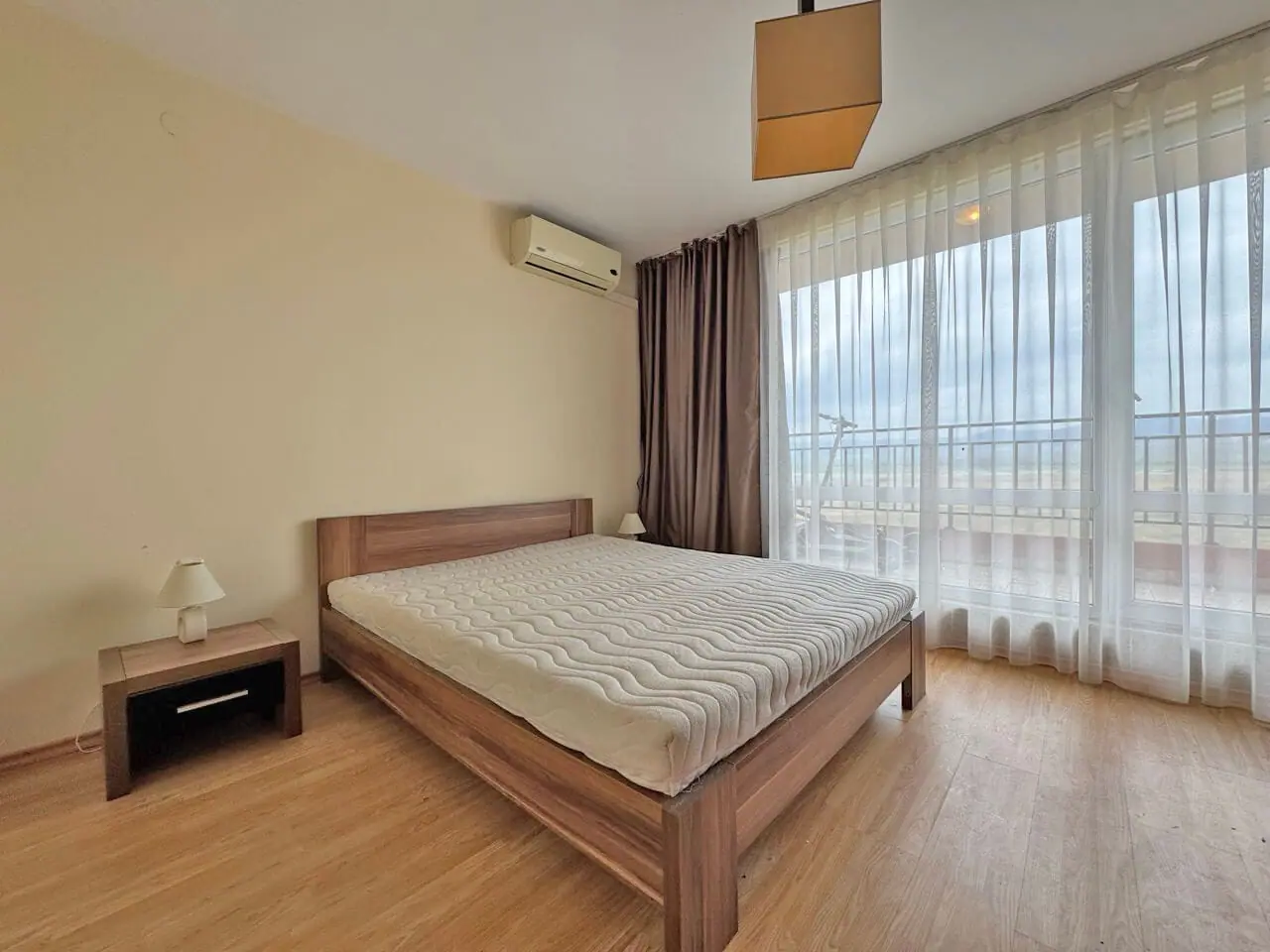 Современная светлая спальня с деревянной кроватью, прикроватной тумбочкой и большими окнами, выходящими на балкон.