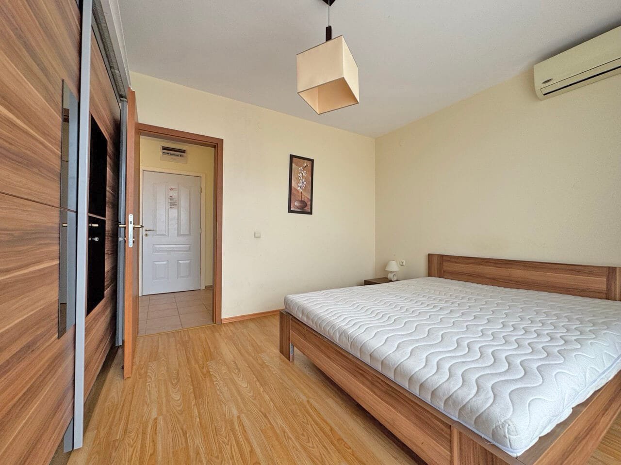 Современная спальня с деревянной мебелью, пустой кроватью и нейтральными стенами, освещенная естественным светом.