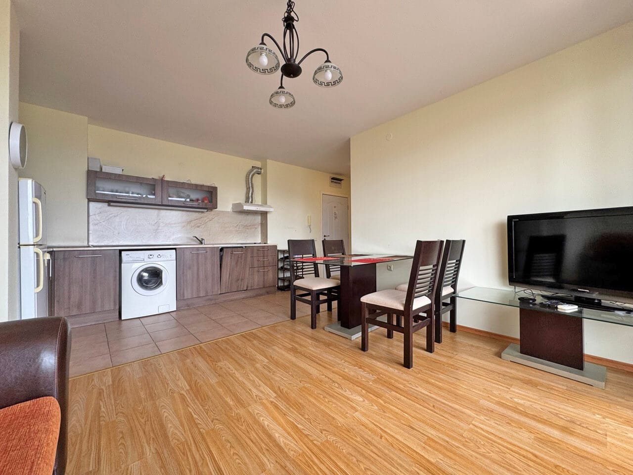 Современная кухня-студия с обеденной зоной, стиральной машиной и телевизором, отделка в нейтральных тонах.