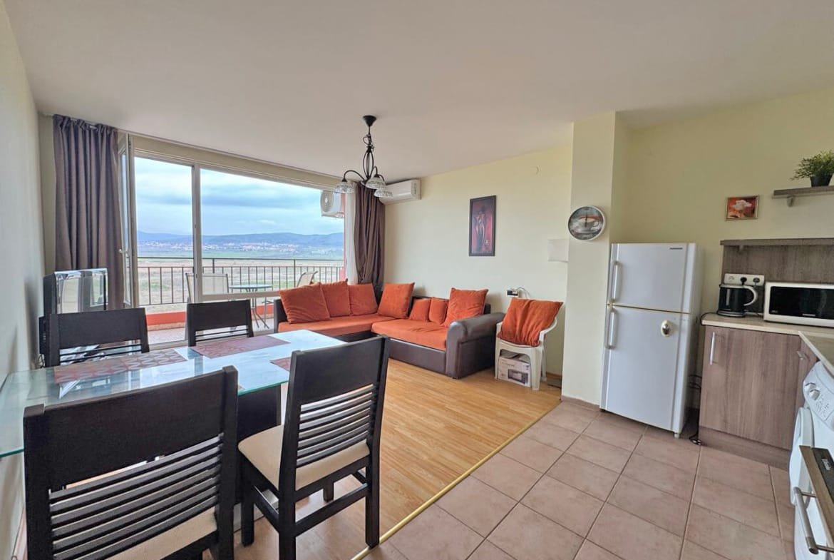 Современная гостиная с оранжевым диваном, обеденным столом и кухонной зоной, большие окна с видом на город.