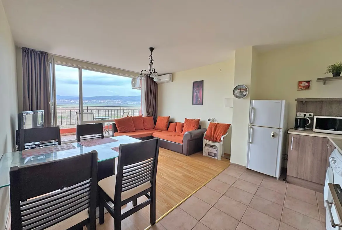 Современная гостиная с оранжевым диваном, обеденным столом и кухонной зоной, большие окна с видом на город.