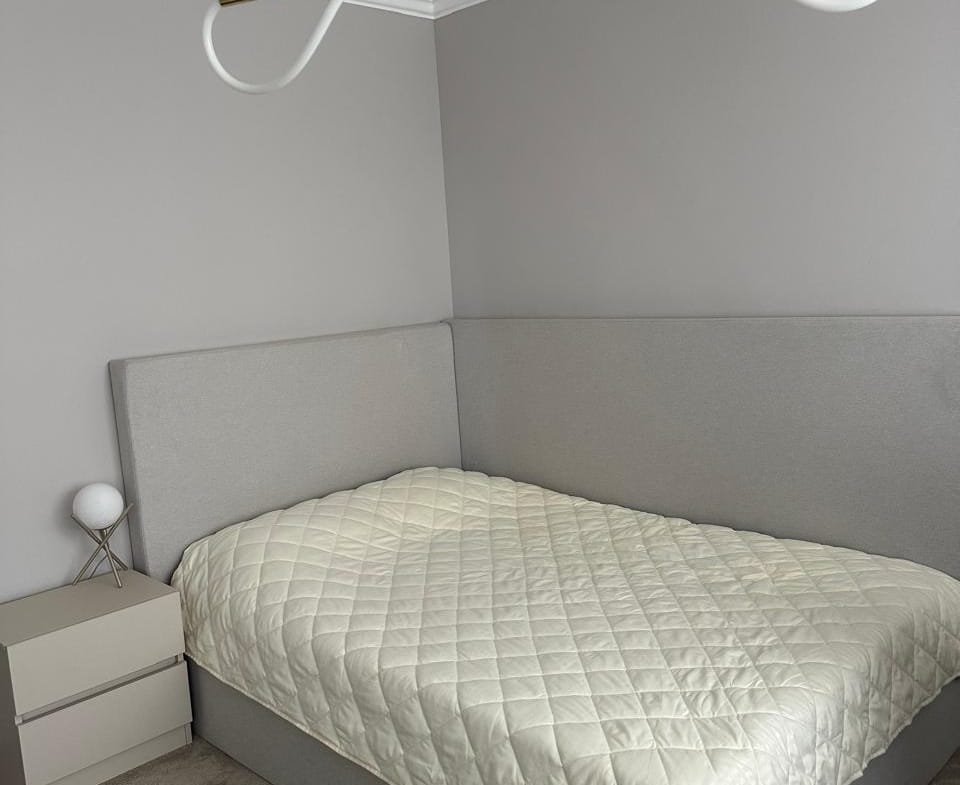 Уютная спальня с серой стеной, большой кроватью и прикроватной тумбой, подходит для покупки недвижимости в Болгарии.