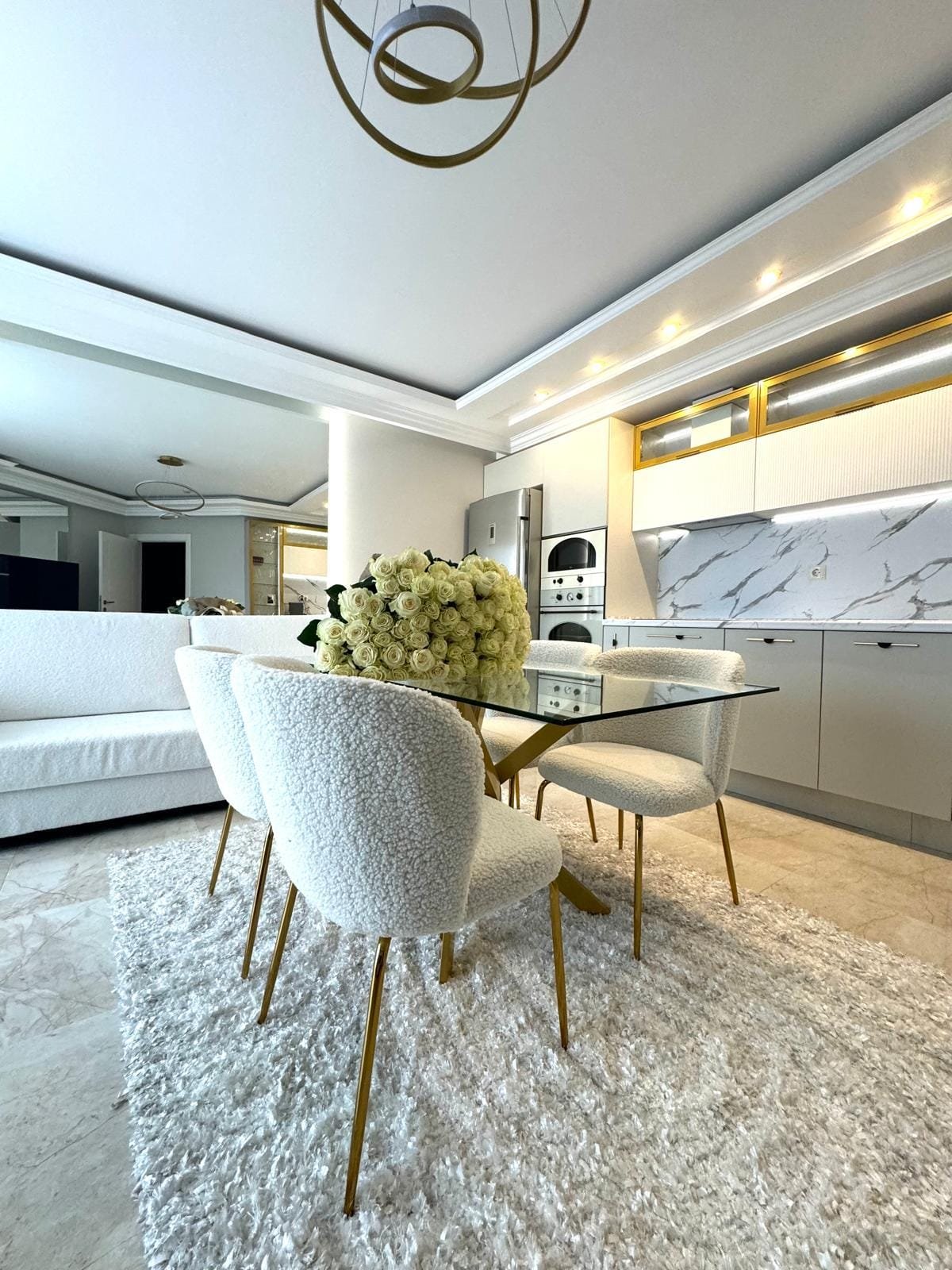 Современная кухня с белыми стульями и круглыми столом, оформленная в нейтральных тонах, с цветами на столе, противопоставляет уют и стиль.