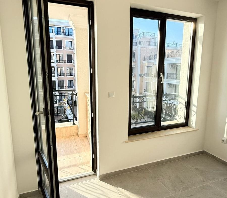 Современная квартира в Болгарии с большими окнами, светлыми стенами и выходом на балкон, идеальная для покупки.