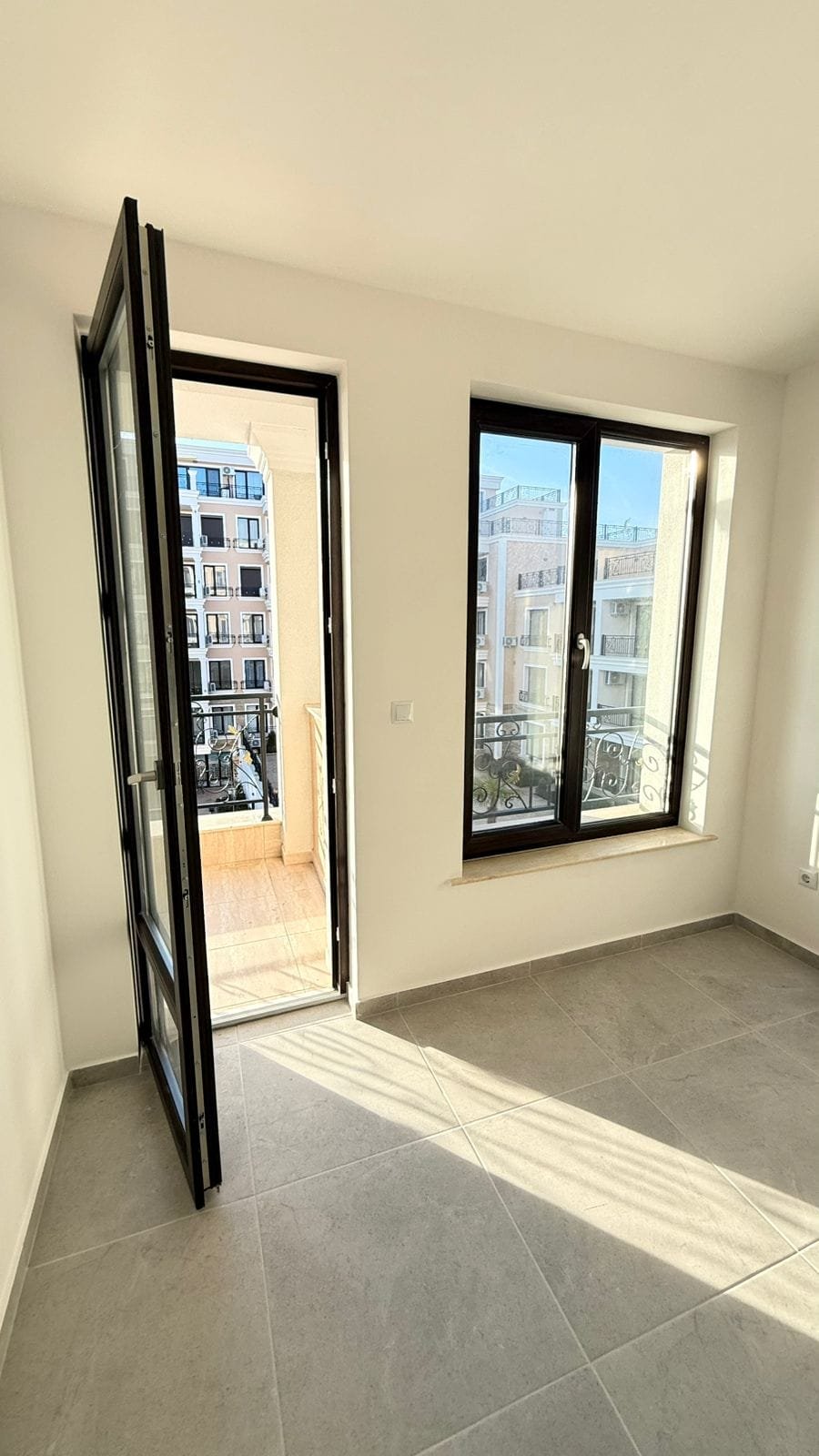 Современная квартира в Болгарии с большими окнами, светлыми стенами и выходом на балкон, идеальная для покупки.