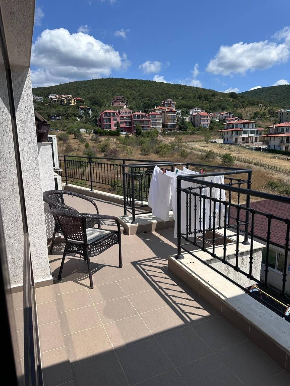 Балкон с плетёной мебелью, развешенное бельё и вид на окружающие дома в курортной зоне Болгарии.