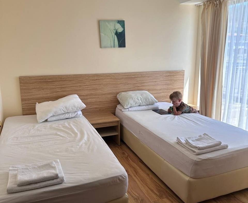 Современная комната с двумя односпальными кроватями, светлыми стенами и большим окном, подходящая для аренды в Болгарии.