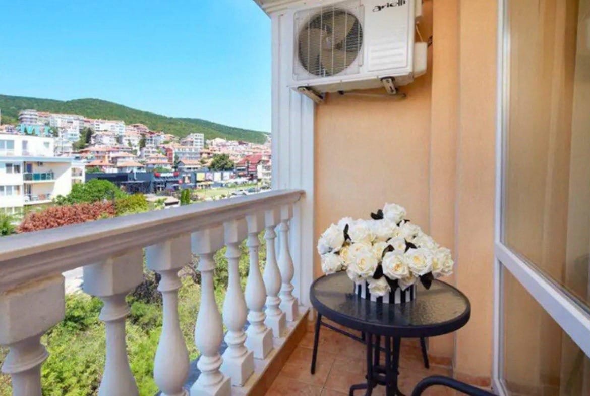Балкон с черным столом и вазой белых роз, виды на жилье и горы, акцент на продаже недвижимости в Болгарии.