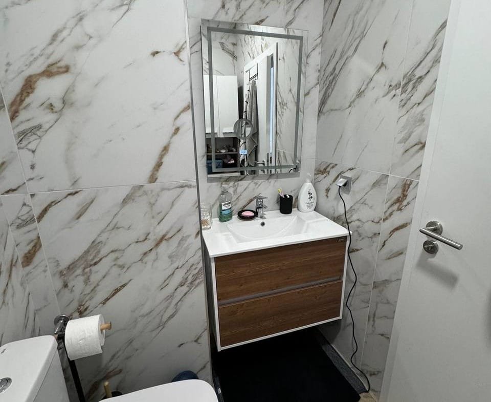 Современный санузел с мраморными стенами, раковиной на деревянной тумбе и зеркалом, удобный для жизни в Болгарии.